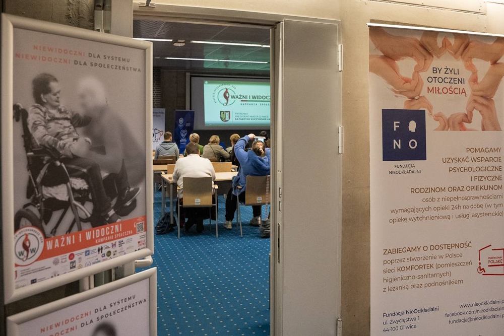 źródło: Fundacja Nieodkładalni/Jadwiga Janowska Zza uchylonych drzwi widać grupę ludzi siedzących przy stołach. Przed wejściem do sali plakaty kampanii "Ważni i widoczni" oraz Fundacji Nieodkładalni
