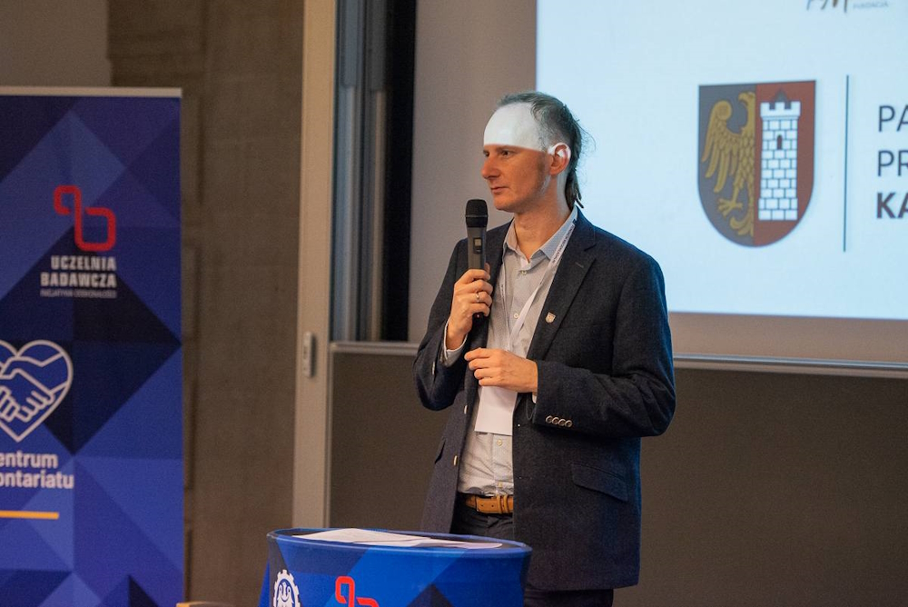 źródło: Fundacja Nieodkładalni/Jadwiga Janowska Zastępca prezydenta miasta - mężczyzna w jasnej koszuli i granatowej marynarce z mikrofonem w ręku. W tle widoczny herb miasta Gliwice wyświetlany na ekranie.
