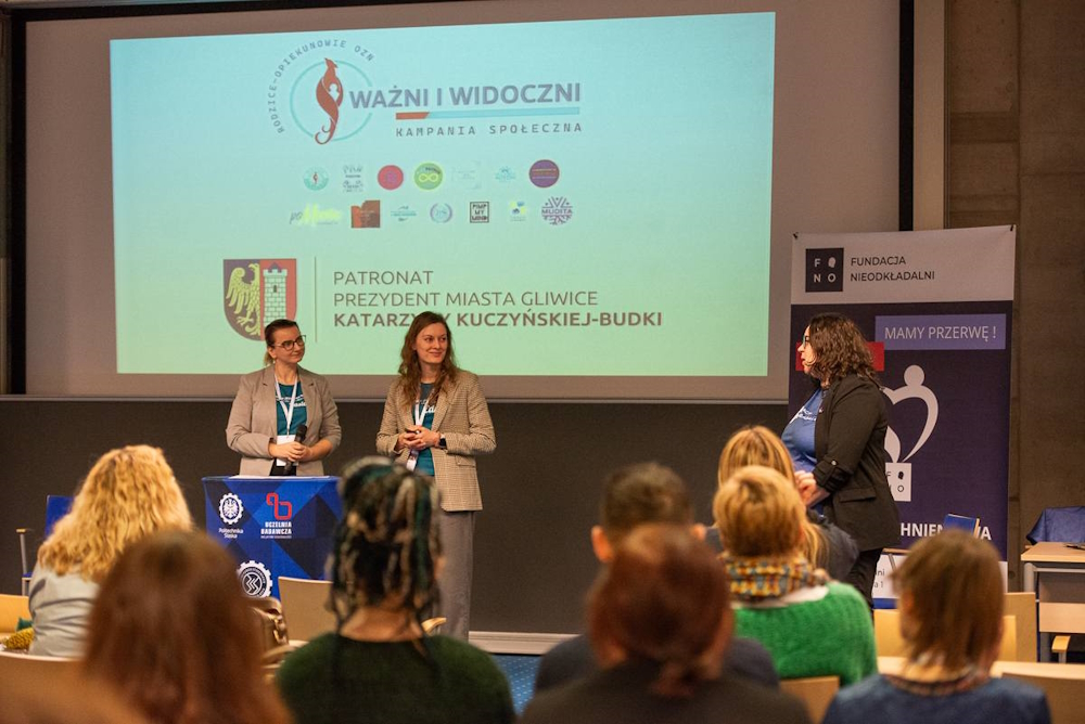 źródło: Fundacja Nieodkładalni/Jadwiga Janowska Dwie kobiety w turkusowych koszulkach i szarych garniturach za mównicą, na tle ekranu. Widoczne także od tyłu kilkanaście osób przysłuchujących się wystąpieniu.