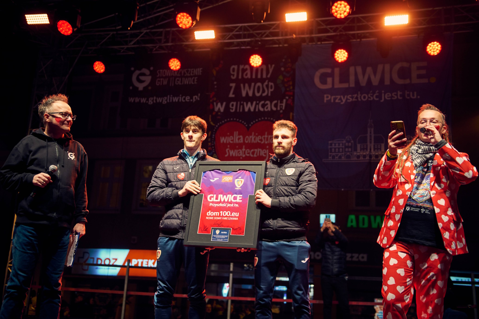 Fot. M. Buksa/UM w Gliwicach Mężczyzna w czerwonej marynarce z serduszami i trzech innych mężczyzn na scenie, dwóch trzyma koszulkę przeznaczoną na licytację