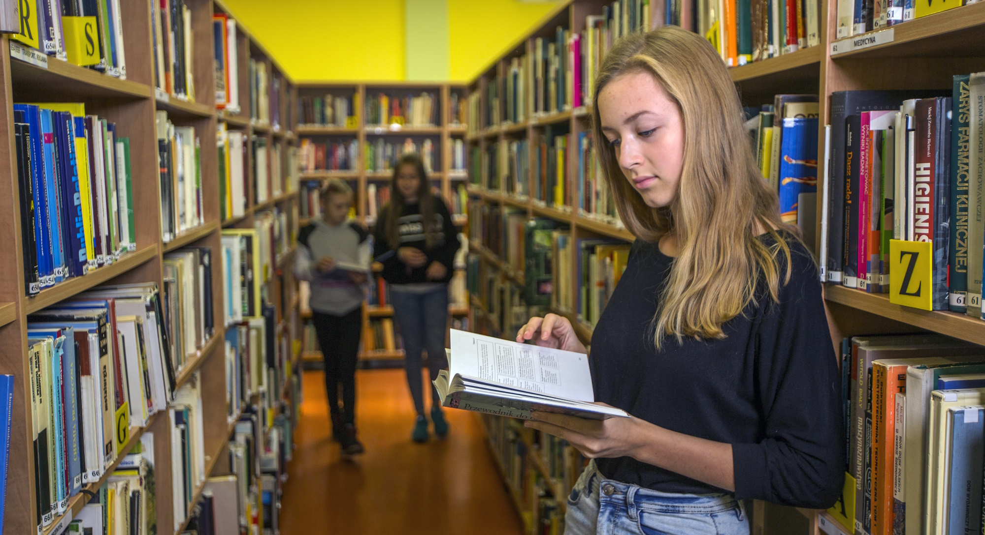 Dziewczynka stoi między regałami w bibliotece i czyta książkę, w oddali widać dwoje innych dzieci