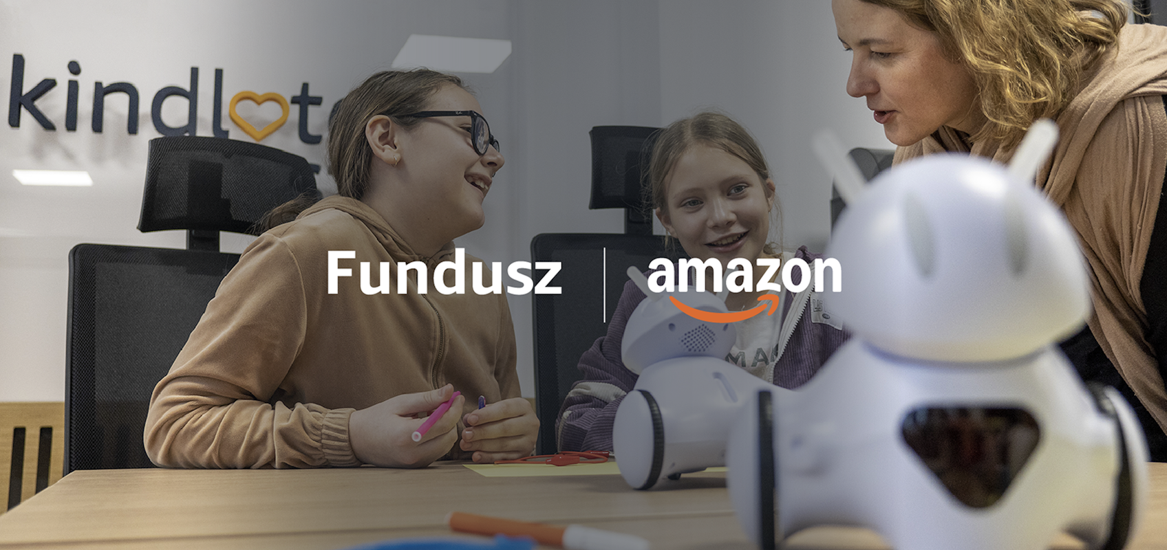 Przy stole siedzą dwie uśmiechnięte dziewczynki, nachyla się nad nimi edukatorka. Na blacie stoją małe roboty.