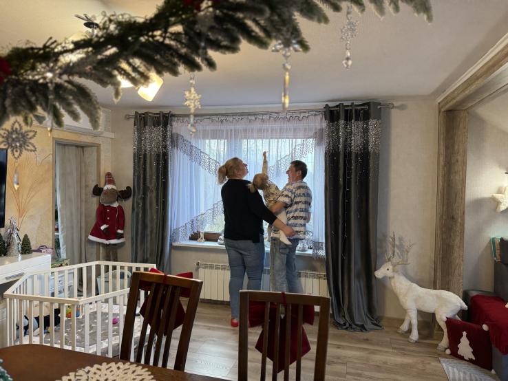 Państwo Miozga z dzieckiem stoją przy oknie w pokoju przystrojonym na święta Bożego Narodzenia